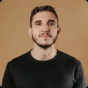 Headshot of a young man with short dark hair and a beard, wearing a black shirt against a beige background.